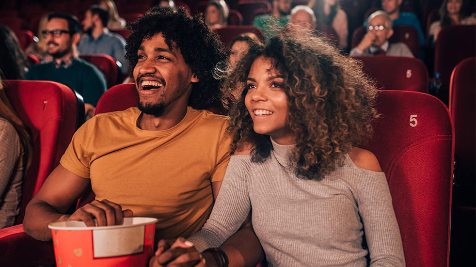 Man and woman holding hands with a big bucket of popcorn, looking happy while watching a film at the cinema